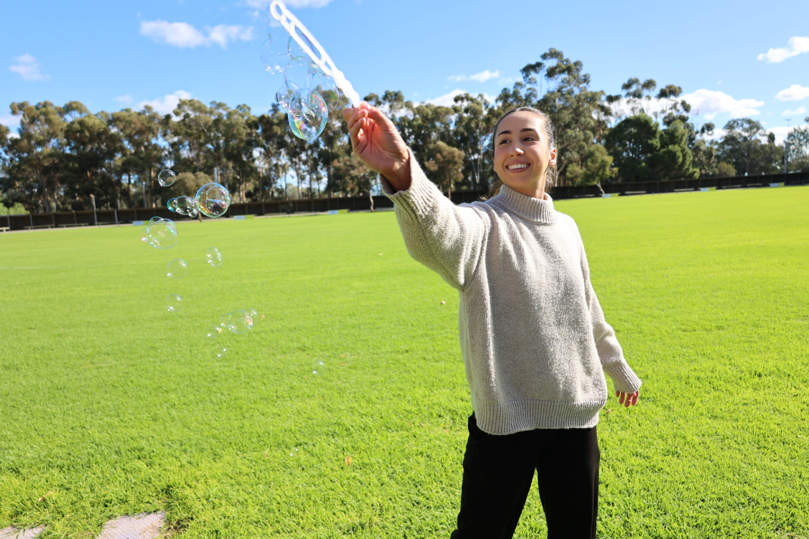 Tamsyn smiling as she blows bubbles across a sunny grassy oval